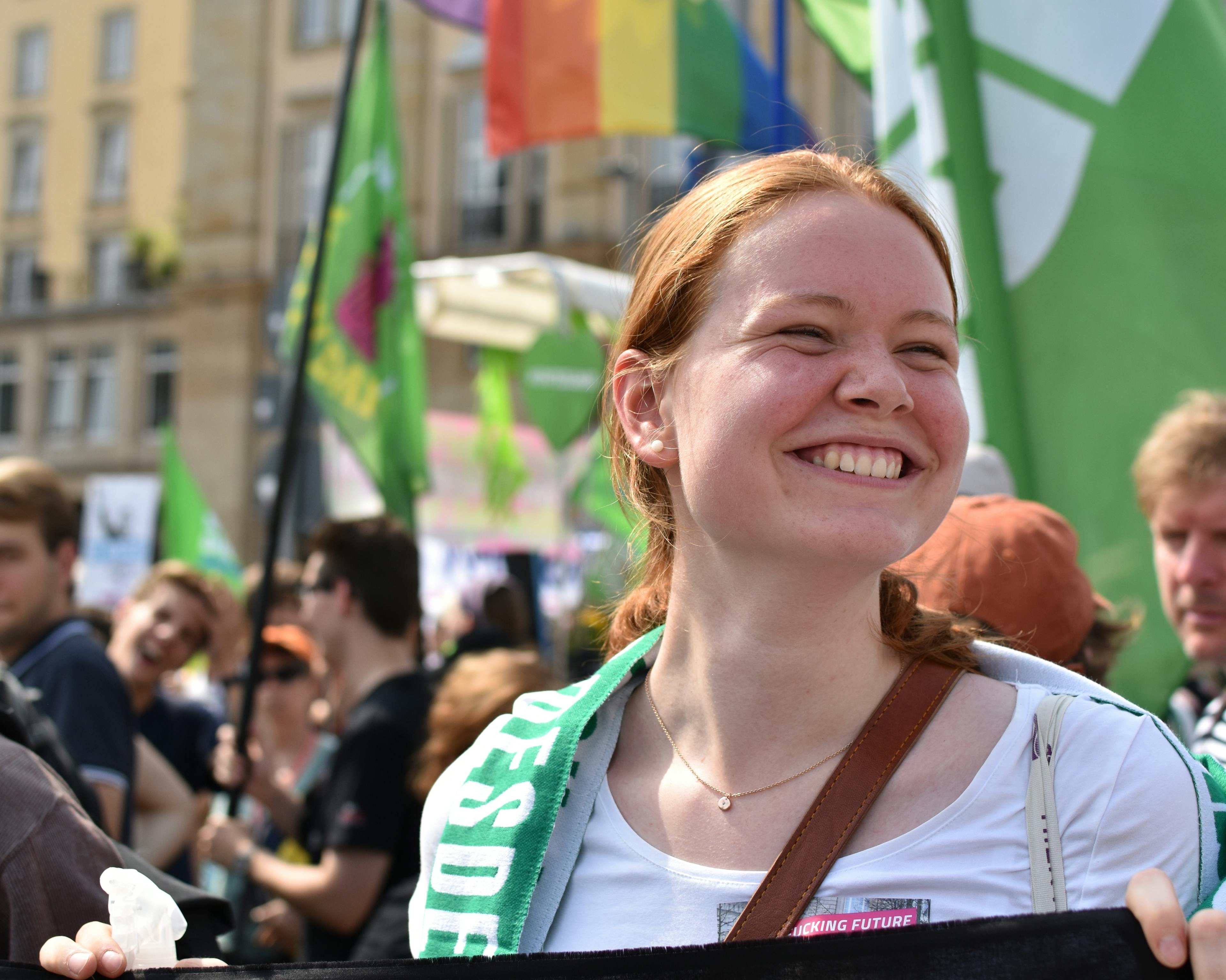 Eine junge Frau hält auf einer Demo ein Banner und strahlt. Im Hintergrund sind viele grüne und bunte Fahnen zu sehen.