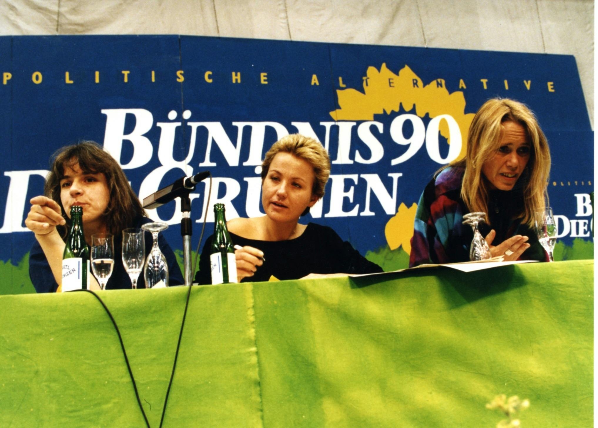 Drei Frauen sitzen an einem Tisch, der mit einem grünen Tuch bedeckt ist und auf dem Mikros und Wasserflaschen stehen. Im Hintergrund ein Schirftzug: "Bündnis 90/DIE GRÜNEN""