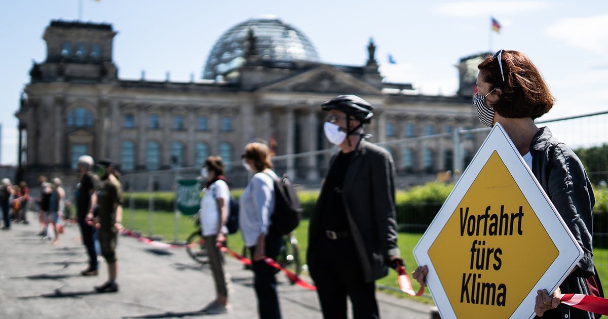Menschen stehen mit Abstand nebeneinander und halten rote Bänder fest. Sie tragen masken. Die Frau im Vordergrund hält ein Plakat in Form eines Verkehrsschildes mit der Aufschrift "Vorfahrt fürs Klima". Im Hintergrund steht der Reichstag.