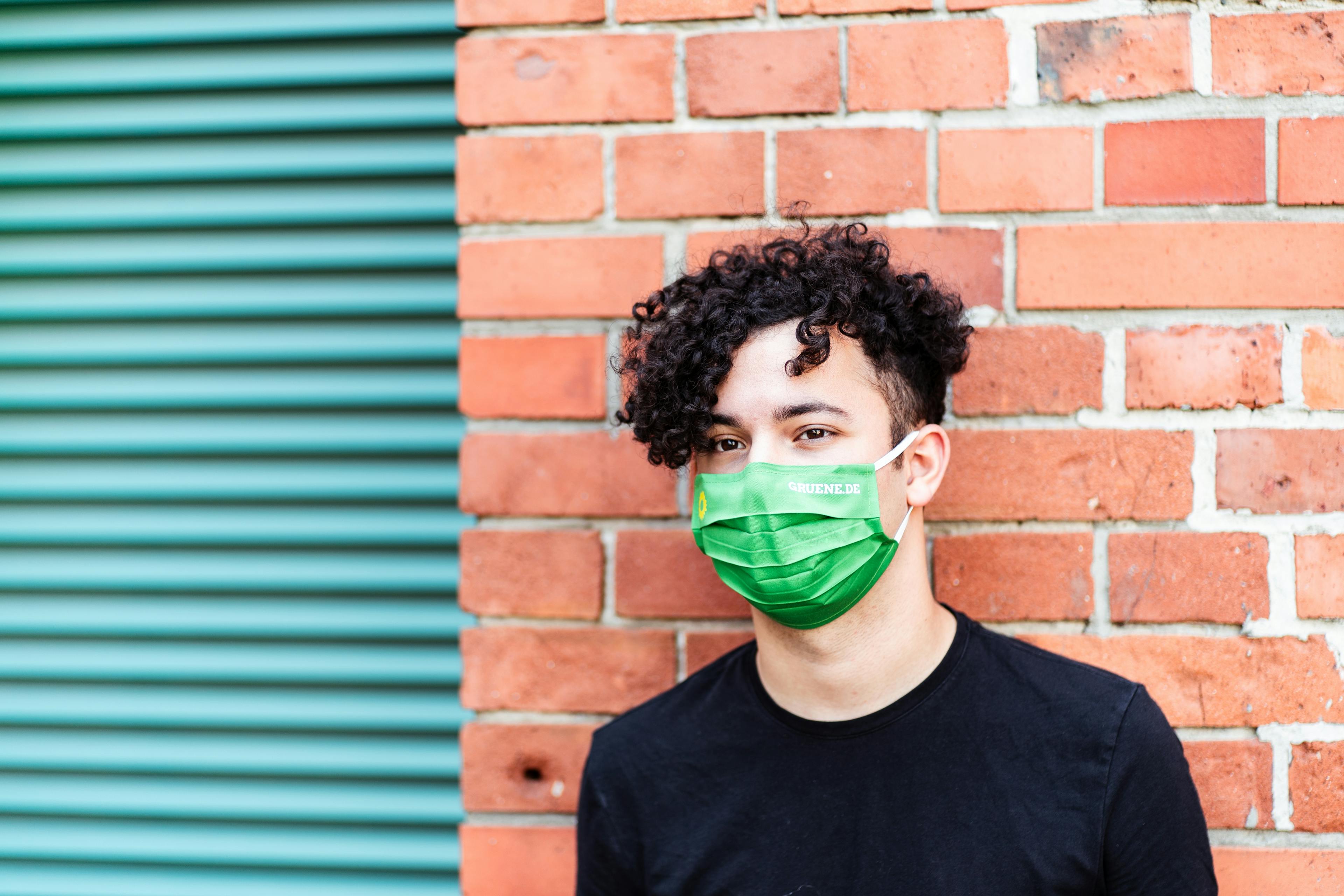 Portrait eines jungen Mannes mit dunklen Locken mit grünem Mund-Nasen-Schutz, der an einer Backsteinmauer lehnt