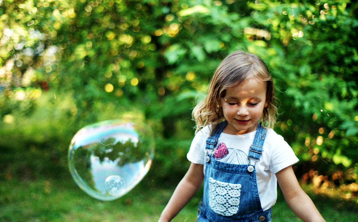 Kleines Mädchen mit Jeans-Latzhose spielt mit einer großen Seifenblase, im Hintergrund sieht man Bäume und eine Wiese