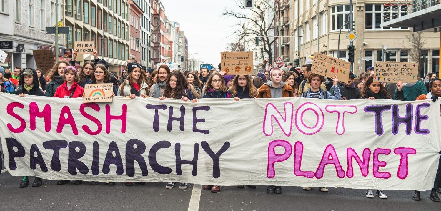 Viele Menschen auf einer Demo tragen ein großes Banner vor sich her, auf dem in pink und lila steht: "Smash the Patriarchy, not the planet"