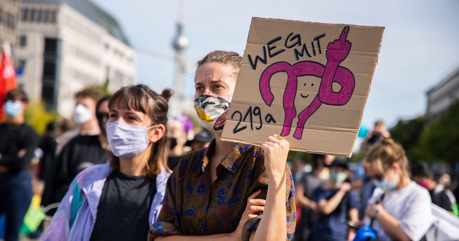 Zwei junge Frauen mit Mund-Nasen-Schutz stehen auf einem Platz. Die eine hält ein Schild mit der Aufschrift "Weg mit 2019a" und der stilisierten Zeichnung von Eierstöcken, die den Mittelfinger zeigen.