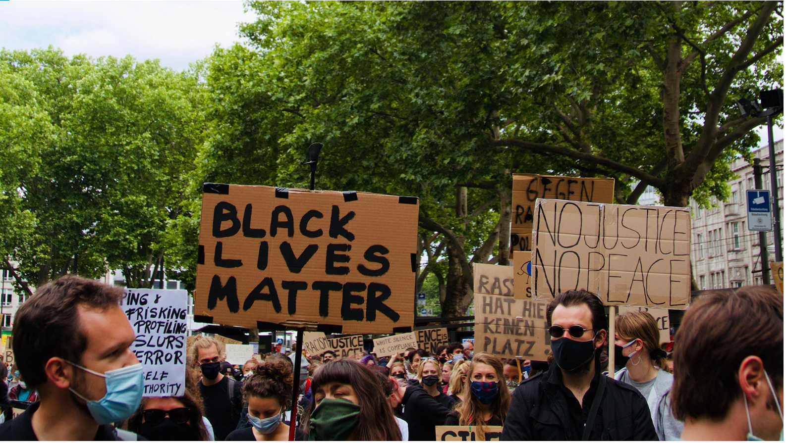 Menschen sind auf einer Demo, sie tragen Mund-Nase-Schutz. In der MItte ein Schild: "Black Lives Matter", rechts ein Schild: "No Justice No Peace"