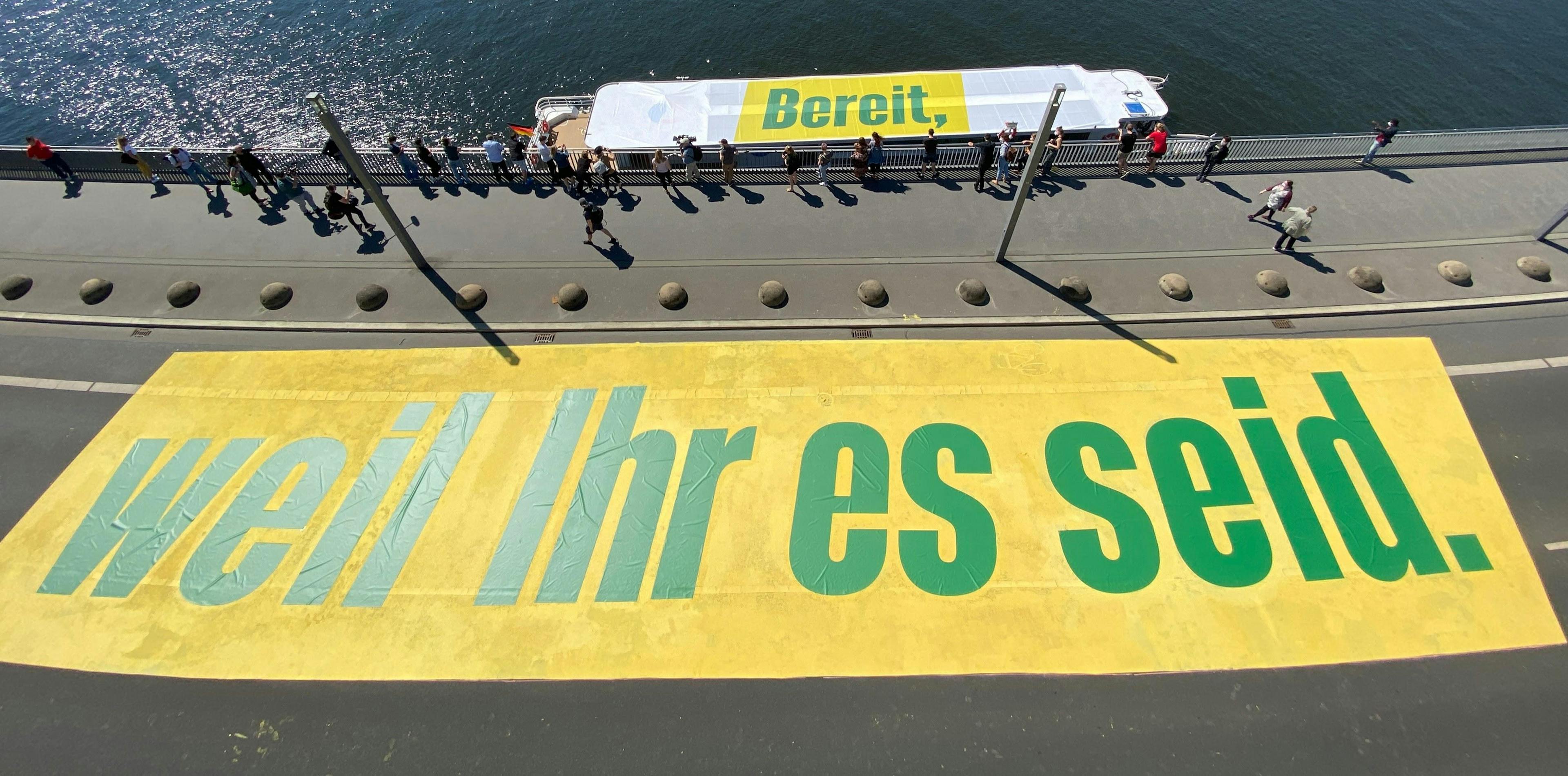 Ein Schiff steht parallel zu einer Brücke. Auf dem Schiff steht in grünen Buchstaben auf gelbem Hintergrund: "Bereit,", auf der Brücke in den selben Farben steht "weil Ihr es seid."