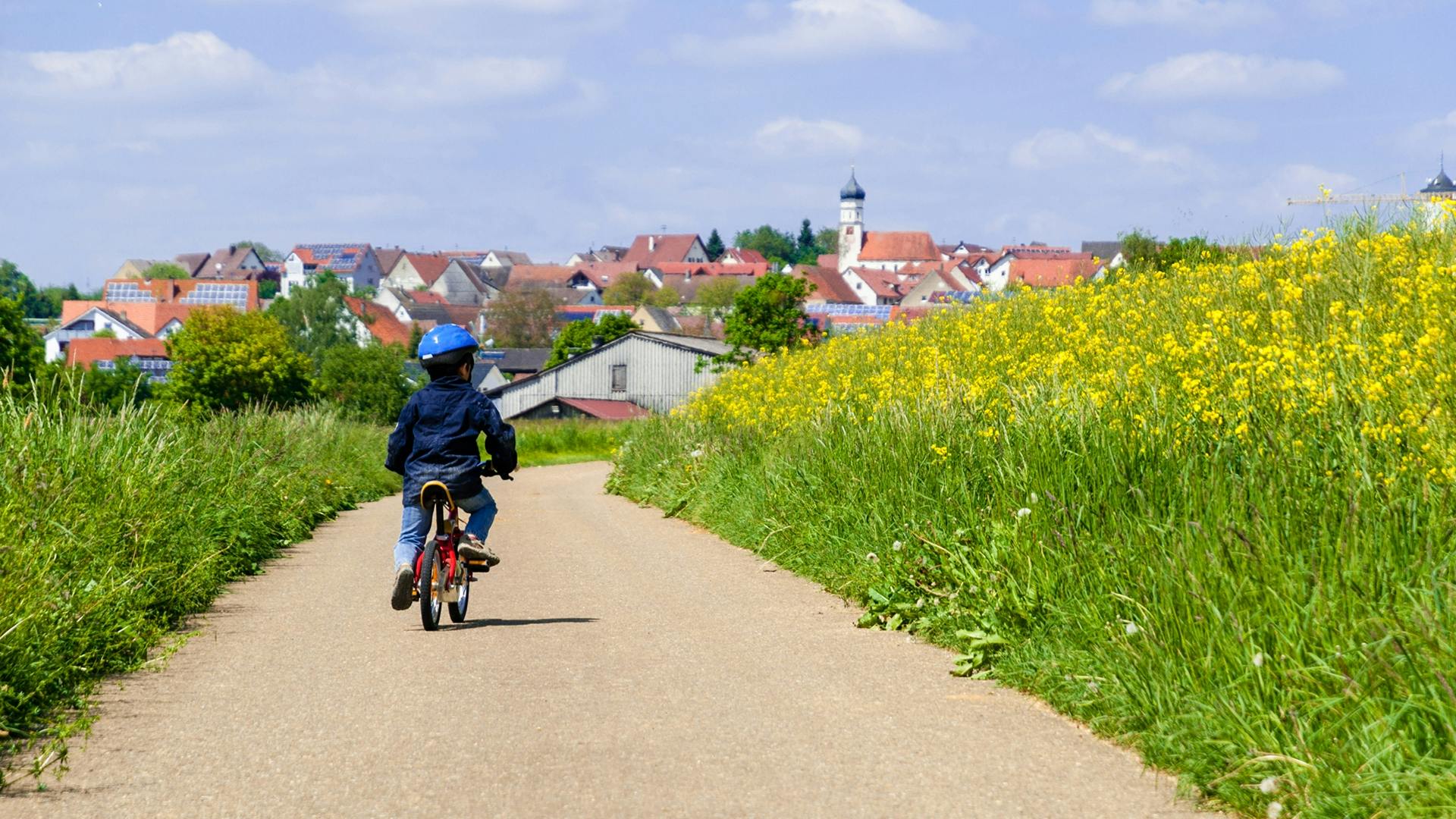Ein Kind fährt auf einem Fahrrad auf einer Straße durch Rapsfelder, die zu einem Dorf führt.