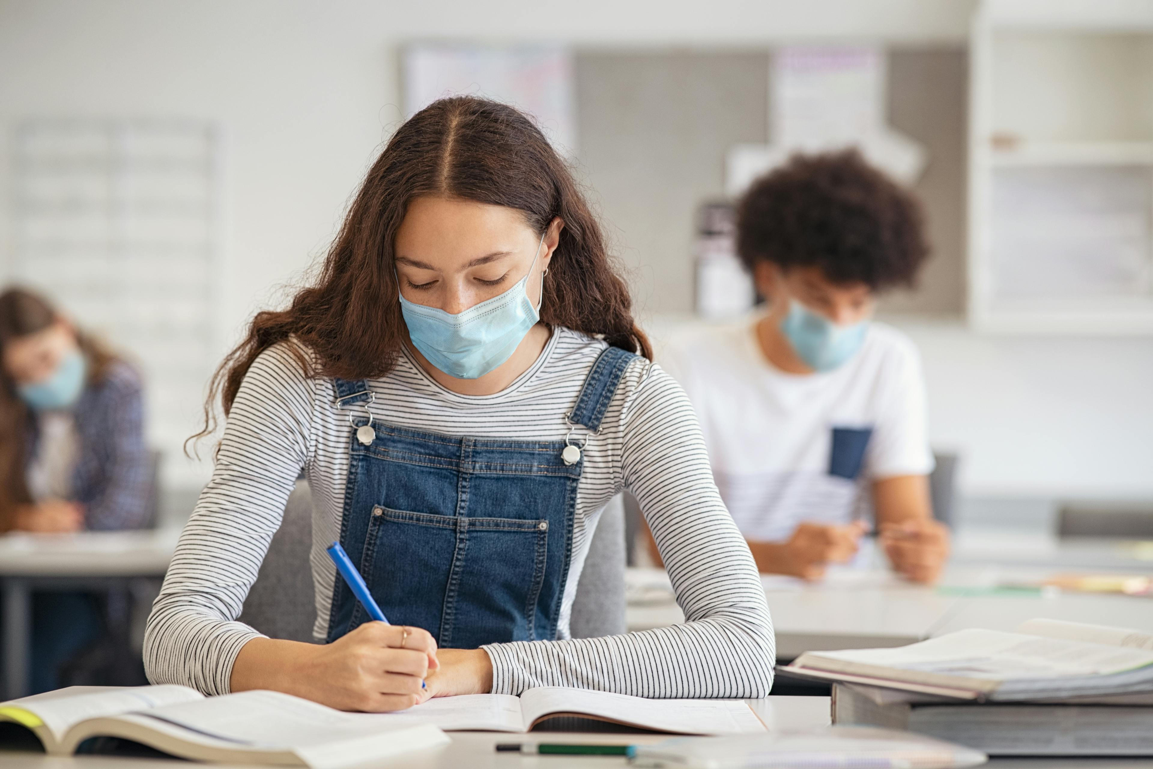 Mädchen mit medizinischem Mund-Nase-Schutz sitzt im Klassenzimmer und schreibt in ihr Heft.