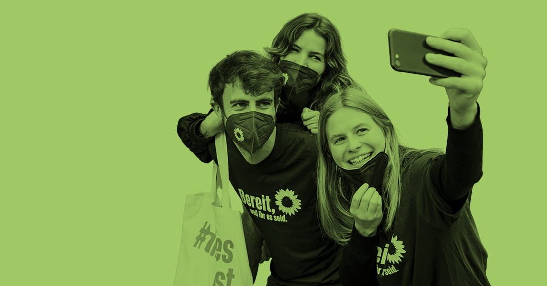 drei junge Menschen machne in Selfie. Der Mann trägt eine der Frauen Huckepack. Alle tragen Mund-Nase-Schutz und T-Shirts der Grünen.