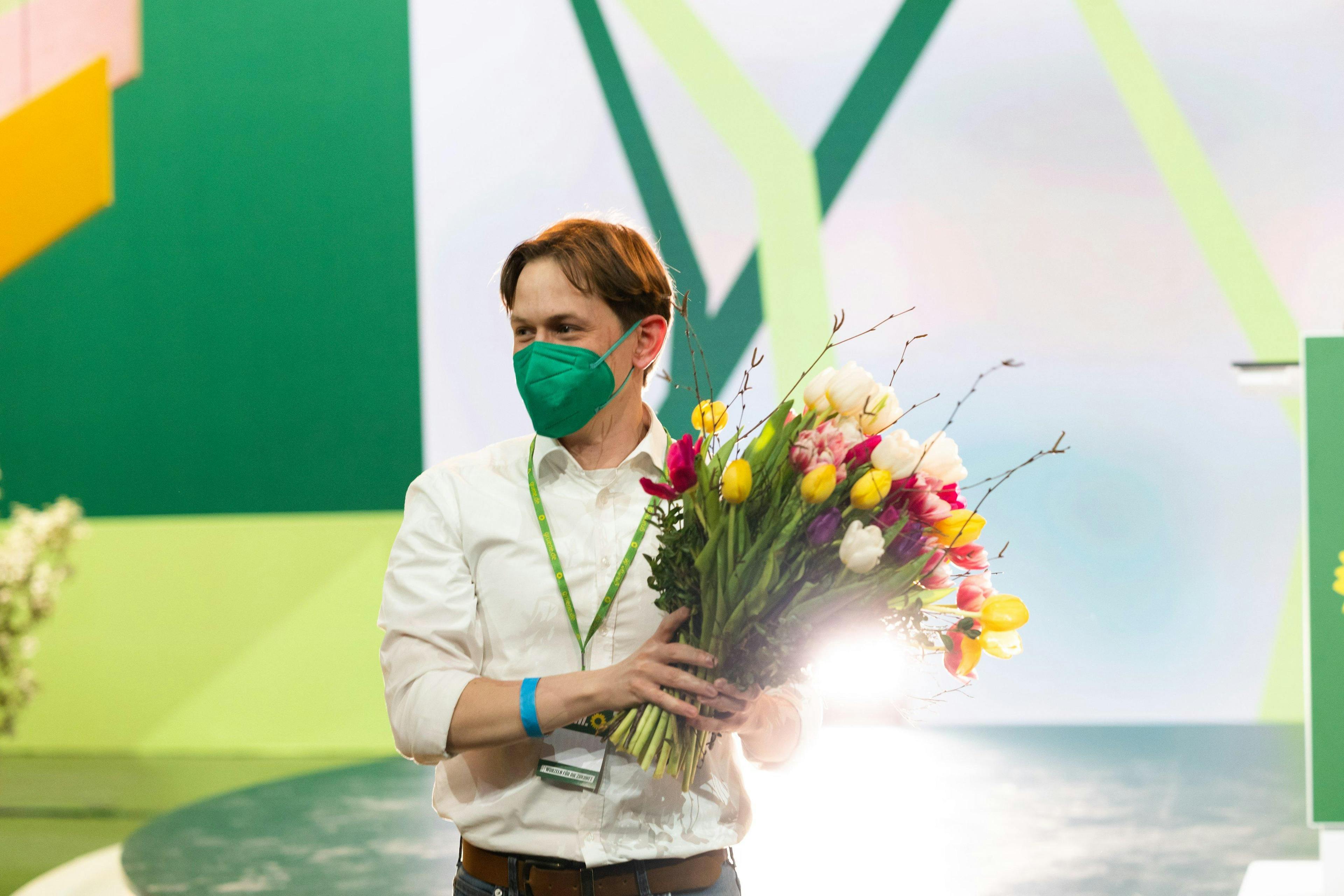 Heiko Knopf steht vor einer Bühne mit grünem Mund-Nase-schutz und hält einen Strauß Tulpen