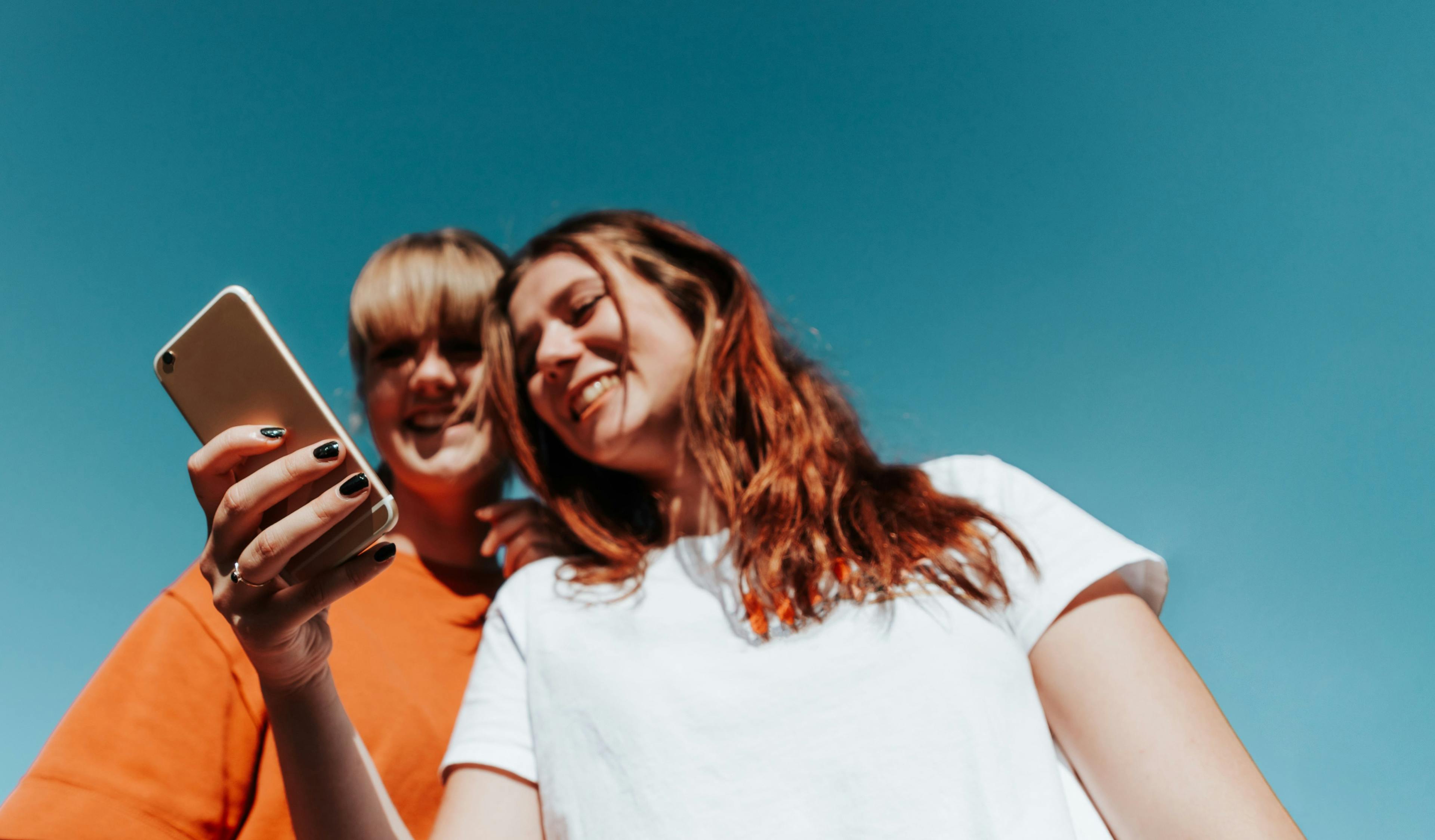 Zwei junge Frauen stehen nebeneinander und schauen auf ein Handy, das die eine in der Hand hält. Im Hintergrund blauer Himmel.