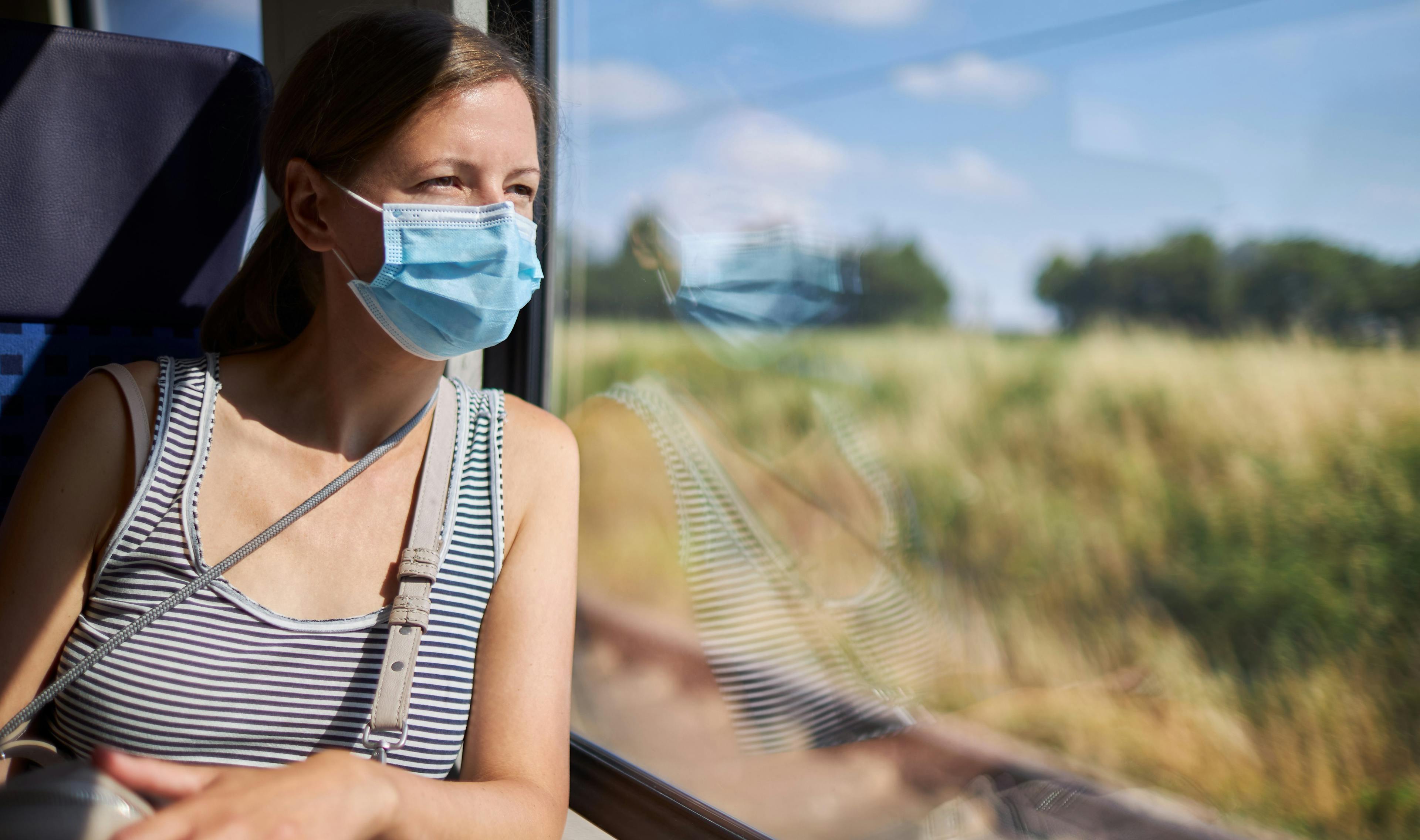 Junge Frau mit gestreiftem Oberteil und medizinischem Mund-Nasenschutz sitzt im Zug und schaut aus dem Fenster.