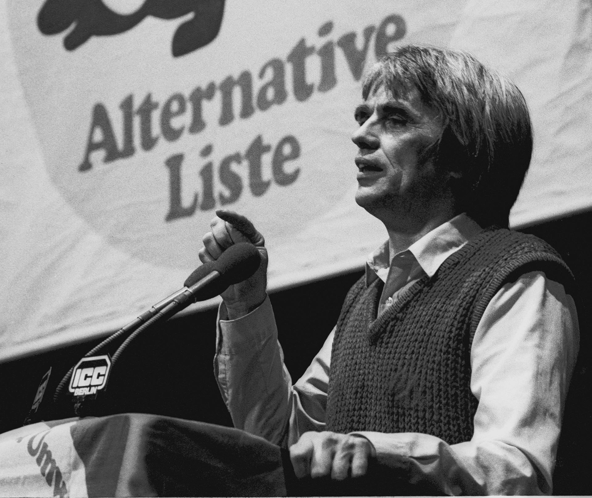 alte Schwarz-Weiß Fotografie von einem jungen Hans-Christian Ströbele, der hinter einem Redner*innenpult steht. Im HIntergrund ein Banner mit der Aufschrift "Alternative Liste"