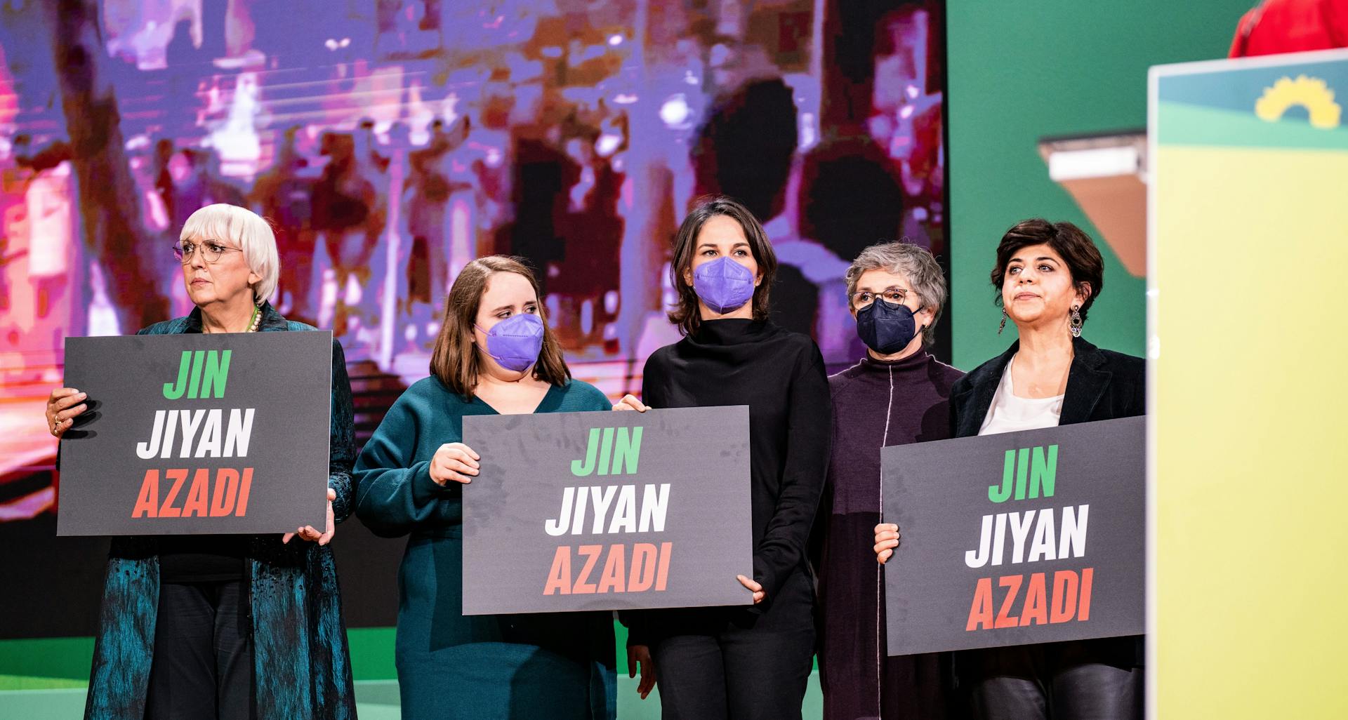 Fünf Frauen stehen nebeneinander vor einer Leinwand, auf der schemenhaft in grellem Lila Menschen zu sehen sind,und halten schwarze Schilder hoch. Die Aufschrift in grün, weiß, schwarz: "Jin Jiyan Azadi"